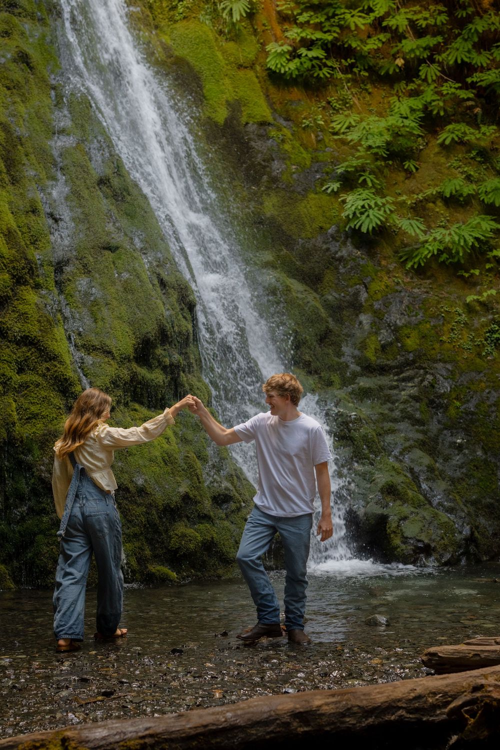 Couple holding hands in front of a lush mossy waterfall, standing in a shallow stream surrounded by green ferns.