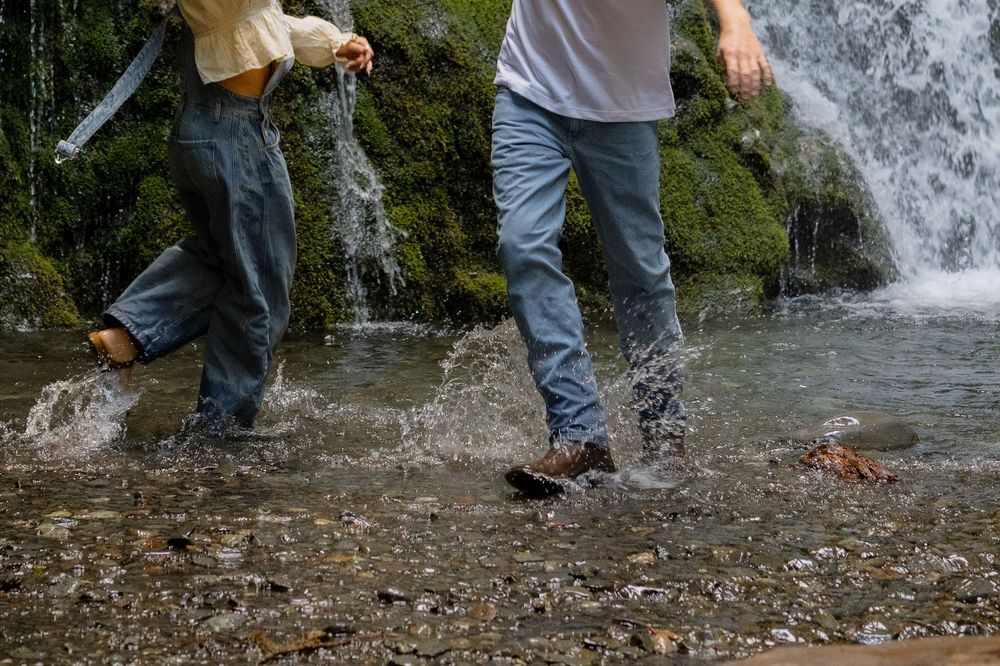 Two people wading barefoot through a rocky stream near a waterfall, splashing water while wearing jeans.