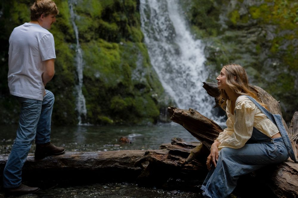 Couple talking near a mossy waterfall with a fallen log in a lush forest setting.