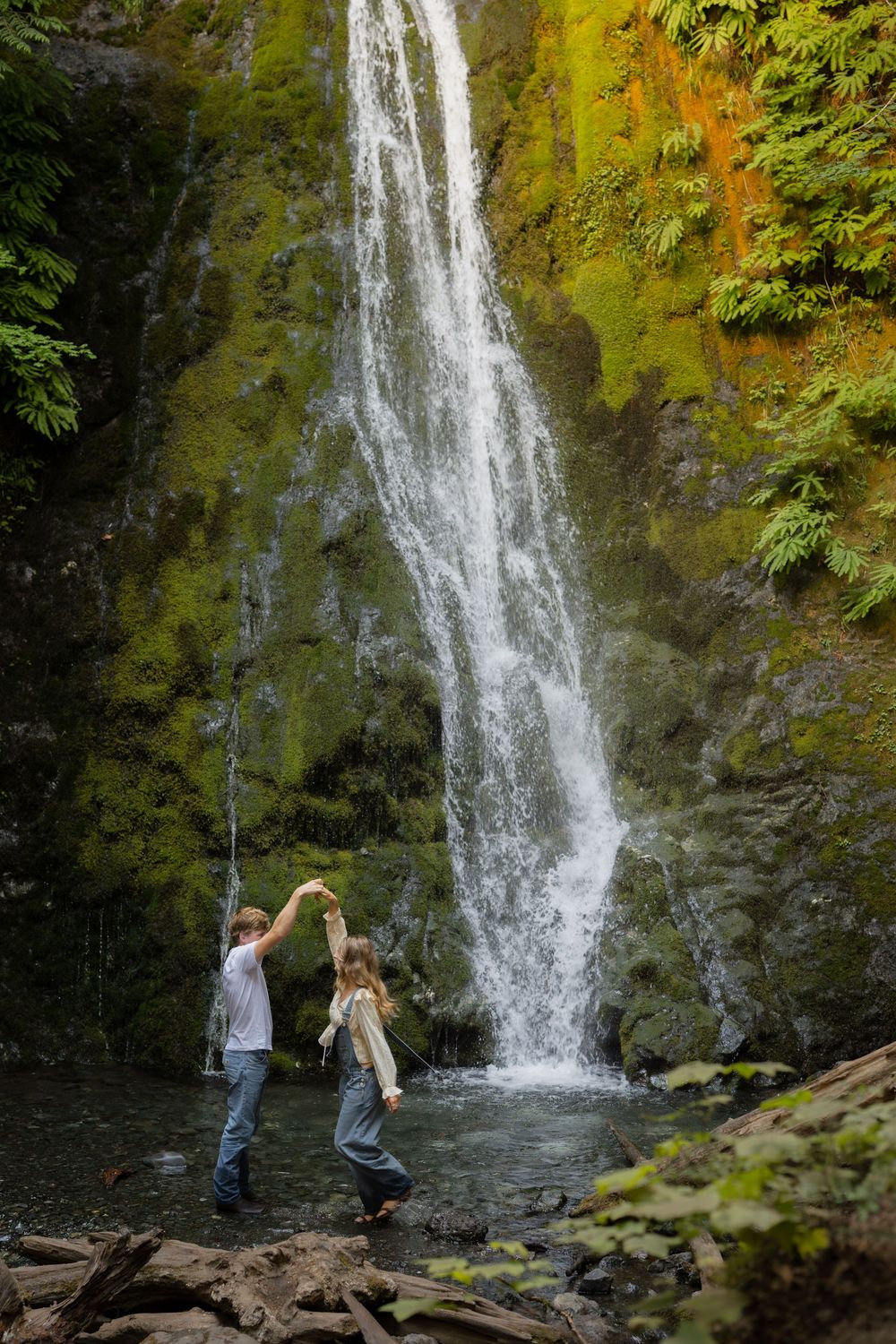 Couple dancing together in front of a tall waterfall surrounded by mossy green cliffs and lush ferns.