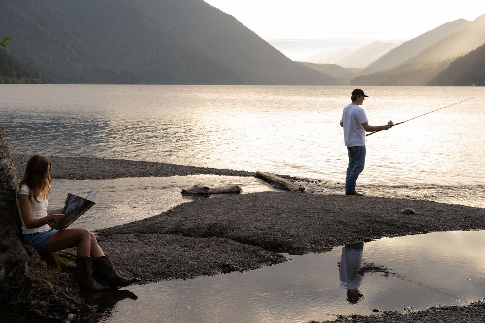Woman reading by a lake at sunset while a man fishes nearby, with mountains reflecting on the calm water.