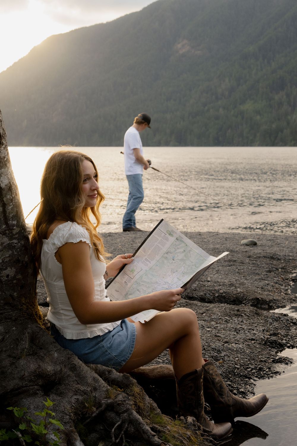 Young woman sits by a lakeside tree reading a map, with a man fishing in the background at golden hour.