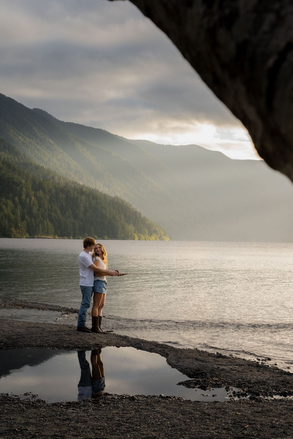 Romantic couple embracing by a misty mountain lake at sunset, reflected in a still puddle.