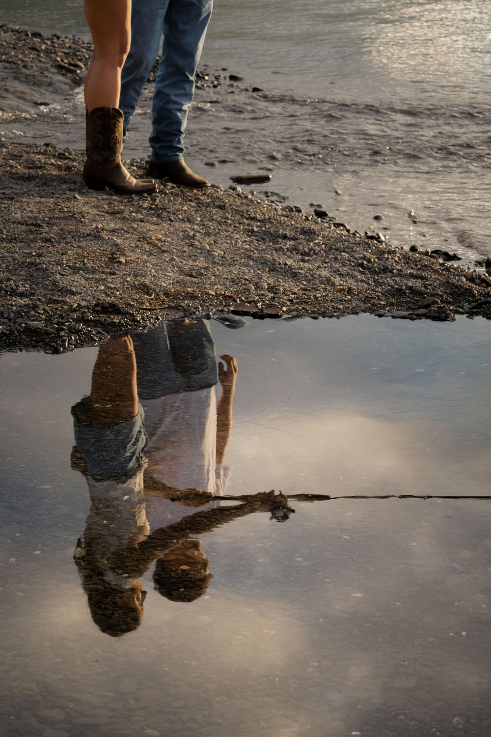 Two people standing on rocky shore reflected in a puddle, wearing jeans and cowboy boots at golden hour.