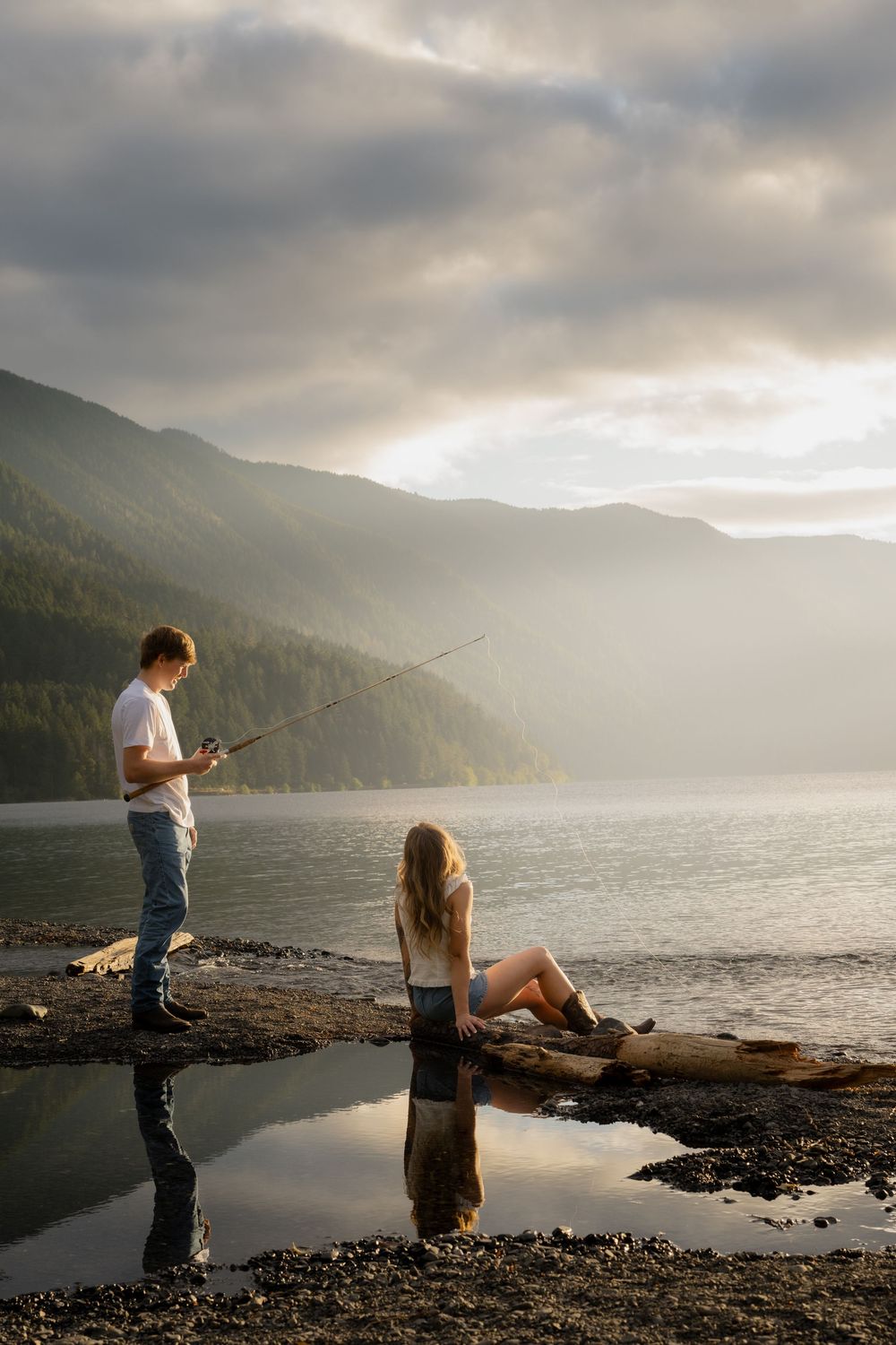 Man fishing with a rod while a woman relaxes on a log by a misty mountain lake at sunset.