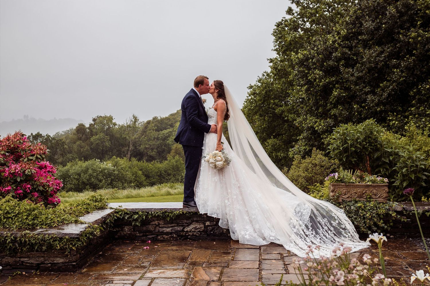 Joyful bride and groom running hand in hand through a vibrant garden with pink rhododendrons on their wedding day.
