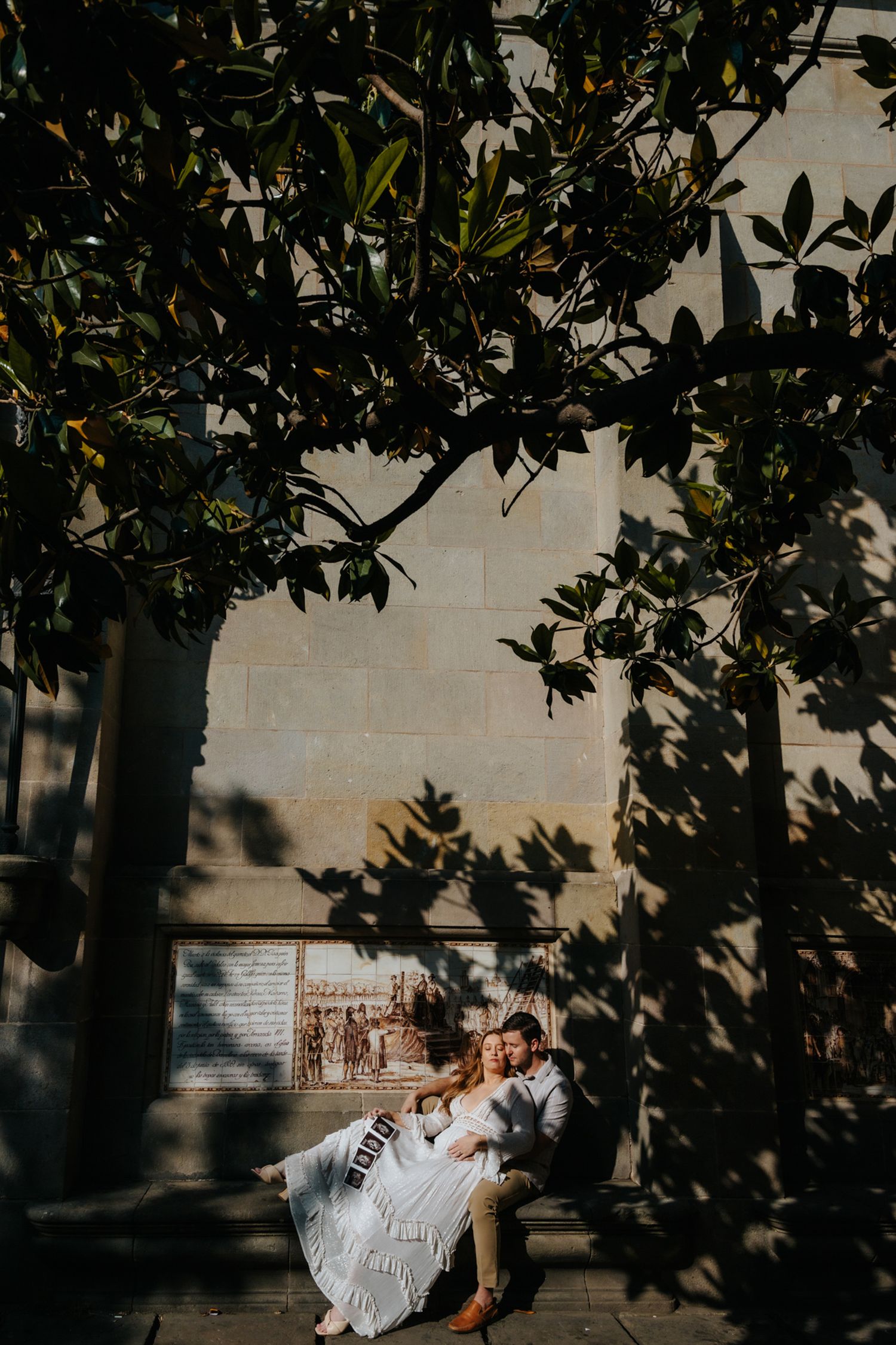 A glowing expectant mother cradling her baby bump while standing under a historic stone archway in Barcelona's Gothic Quarter.