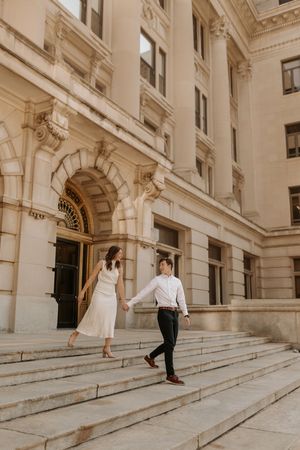 Couple holding hands walking down grand stone steps of elegant beige classical building in wedding attire.