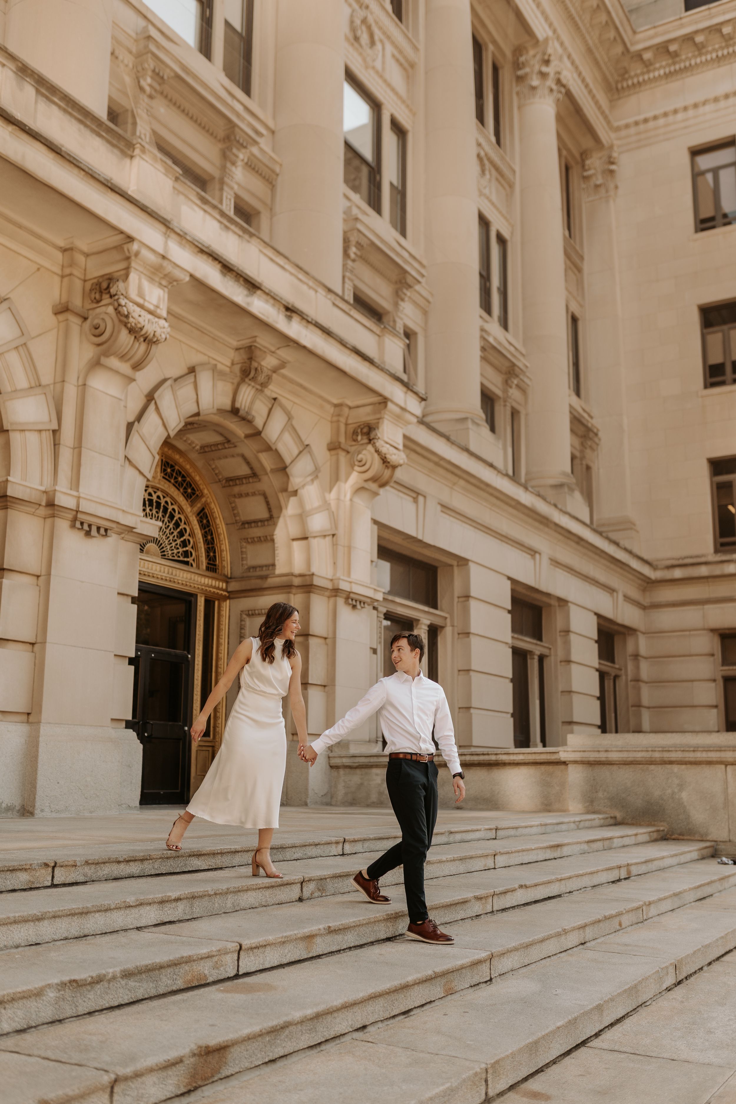 Couple holding hands walking down grand stone steps of elegant beige classical building in wedding attire.
