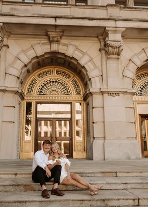 Couple sitting on stone steps outside an ornate building with golden arched doors for an engagement photo.