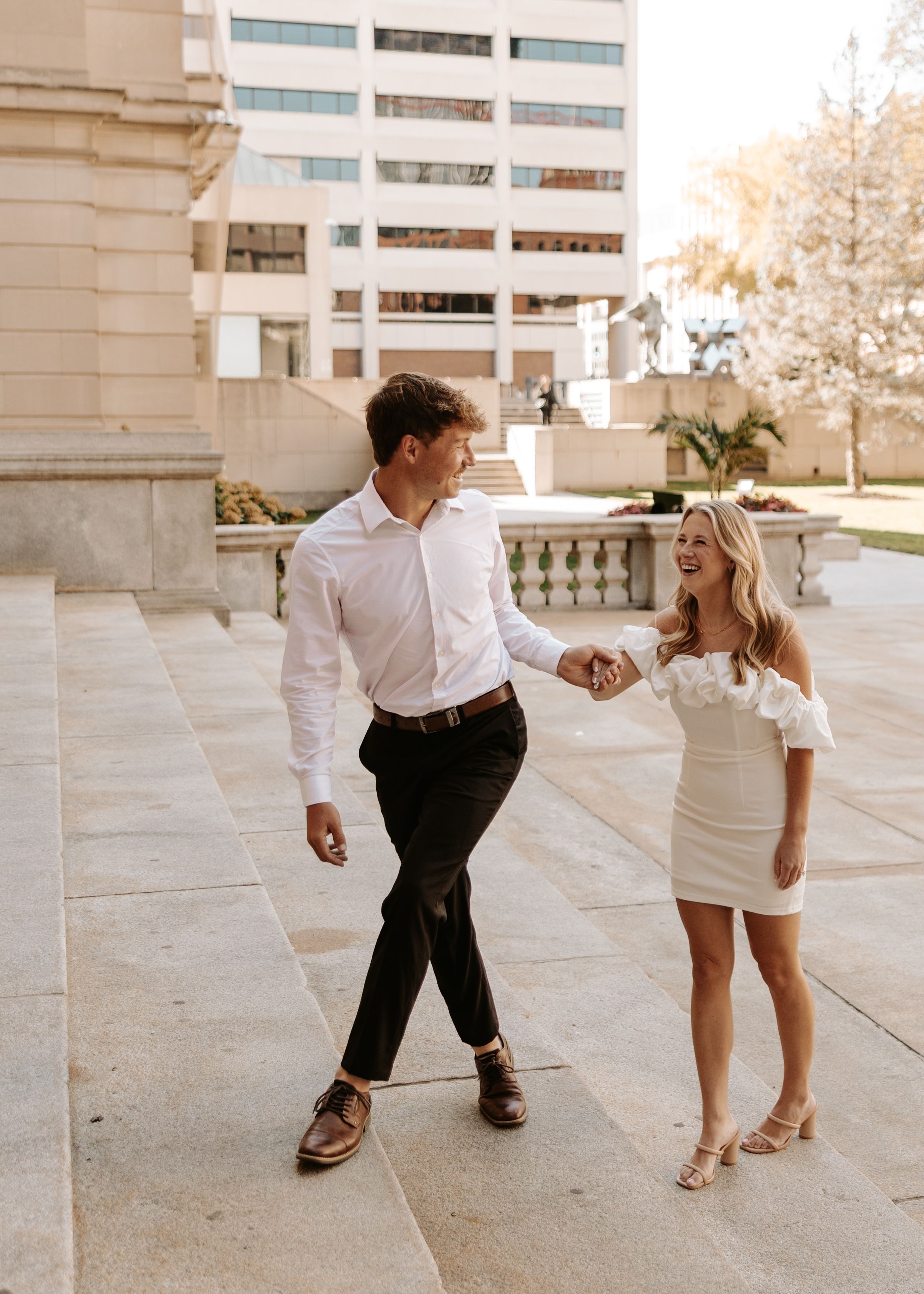 Couple in white outfits holding hands and smiling while walking outdoors near a modern building on a sunny day.