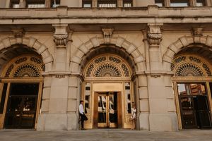 Elegant neoclassical building entrance with ornate arched doorways, gold details, and people standing near revolving doors.
