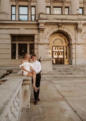 Couple posing on stone steps outside a grand historic building, woman in white romper seated on ledge, man standing beside her.