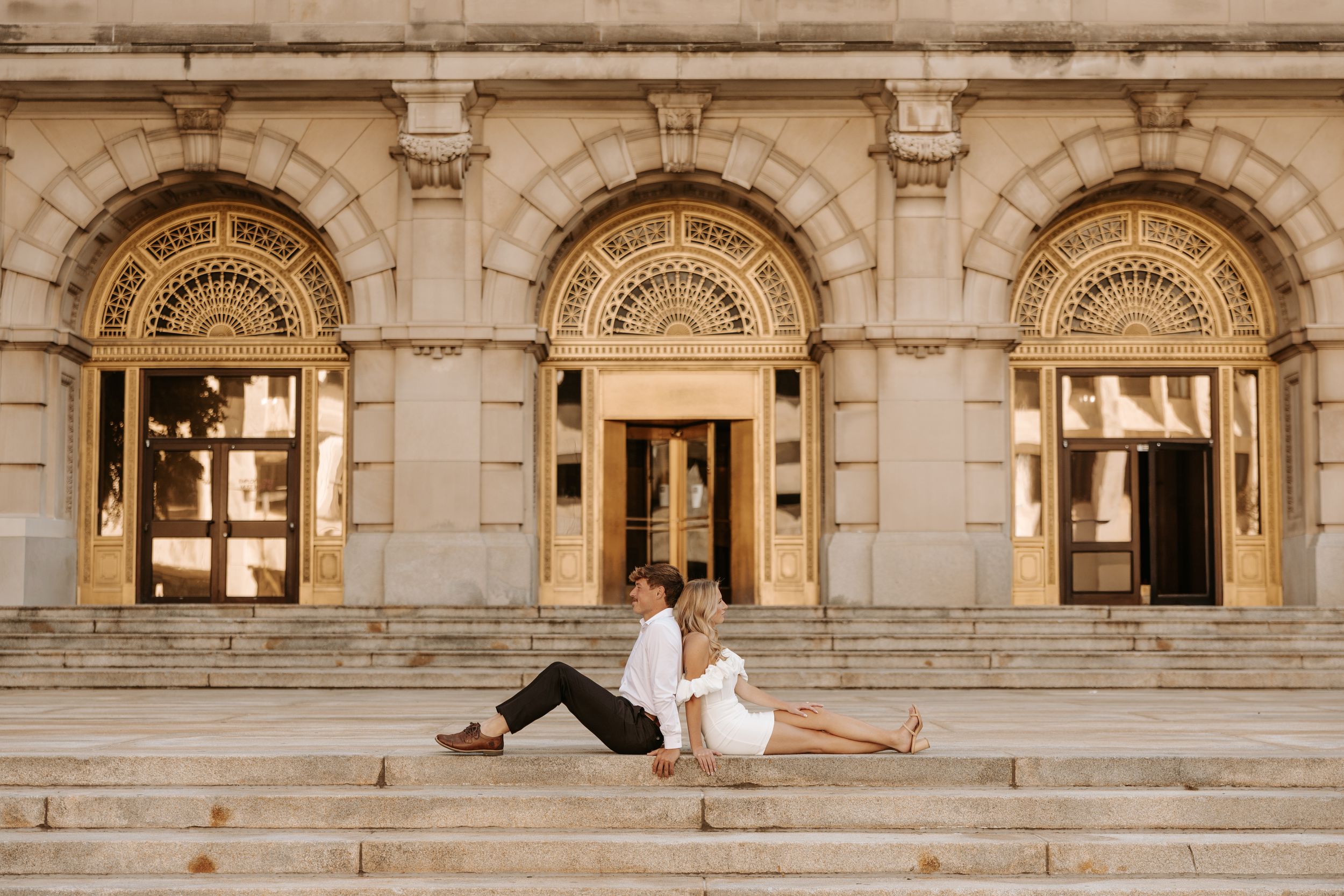 Couple sitting on steps of ornate beige stone building, woman in white dress, man in white shirt, elegant architecture.