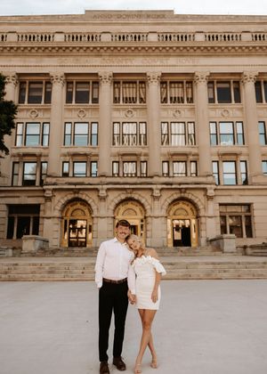 Couple in white outfits posing in front of a grand neoclassical building with arched windows and stone columns.
