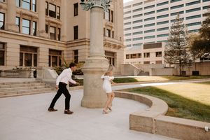 Couple playfully dancing around a ornate stone column in an urban plaza with modern buildings in the background.