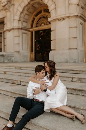 Couple in white sitting on stone steps of elegant building, sharing a romantic moment during engagement photo session.