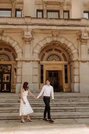 Couple holding hands on steps of ornate historic building, woman in white dress, man in dark pants and white shirt.