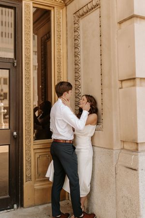 Couple embracing and smiling by ornate gold-framed door, woman in white dress, man in white shirt and gray pants.