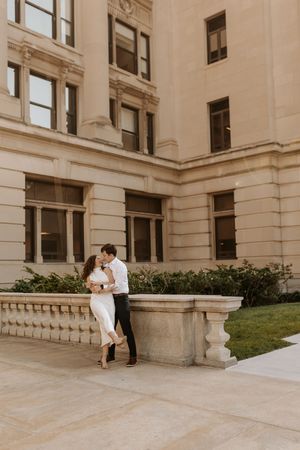 Couple sharing a romantic embrace outside a grand beige stone building with arched windows and manicured hedges.