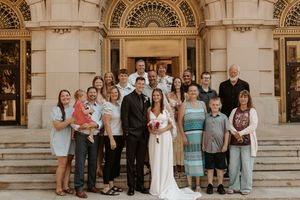 Wedding couple poses with family group on steps of ornate stone building, bride in white gown holding red bouquet.