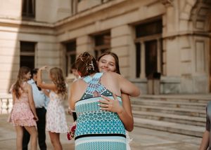 Two young women share a warm hug outside a grand building during a formal event.