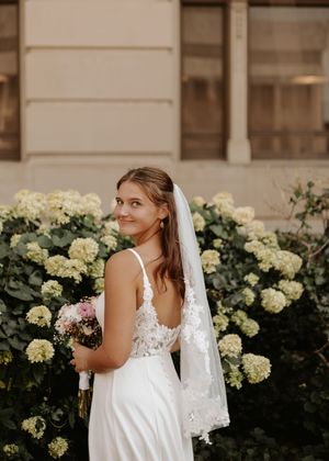 Bride in white lace gown and veil holds bouquet, smiling over shoulder before blooming hydrangea bushes.