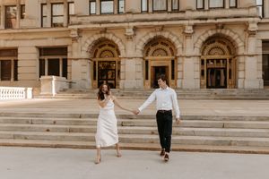 Couple holding hands walking in front of a grand stone building with arched windows and steps.