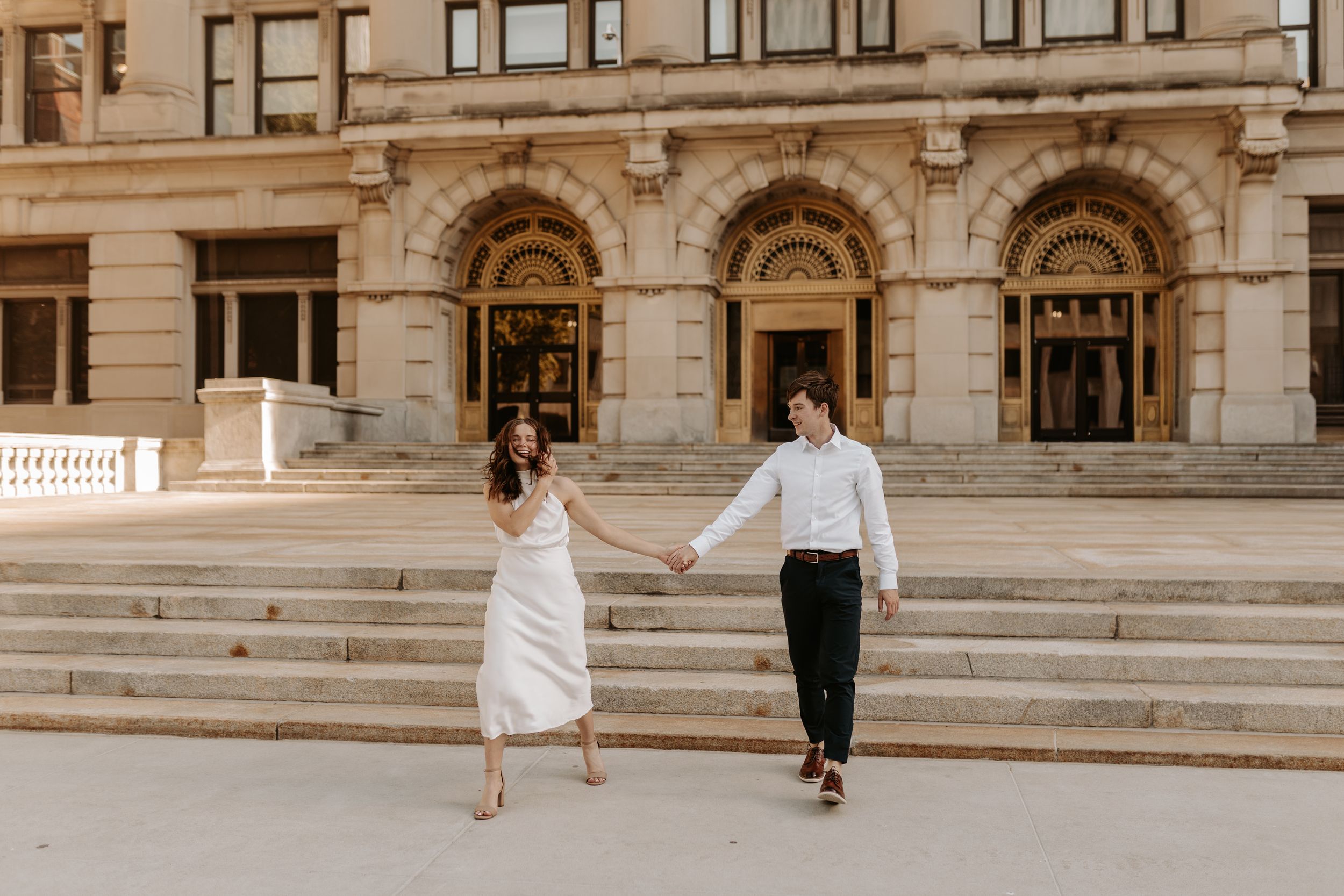 Couple holding hands walking in front of a grand stone building with arched windows and steps.