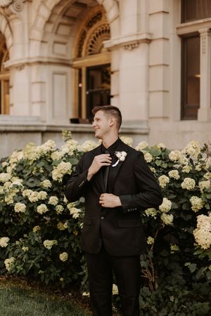 Smiling groom in black suit adjusting boutonniere in front of blooming hydrangea bushes and elegant stone building.