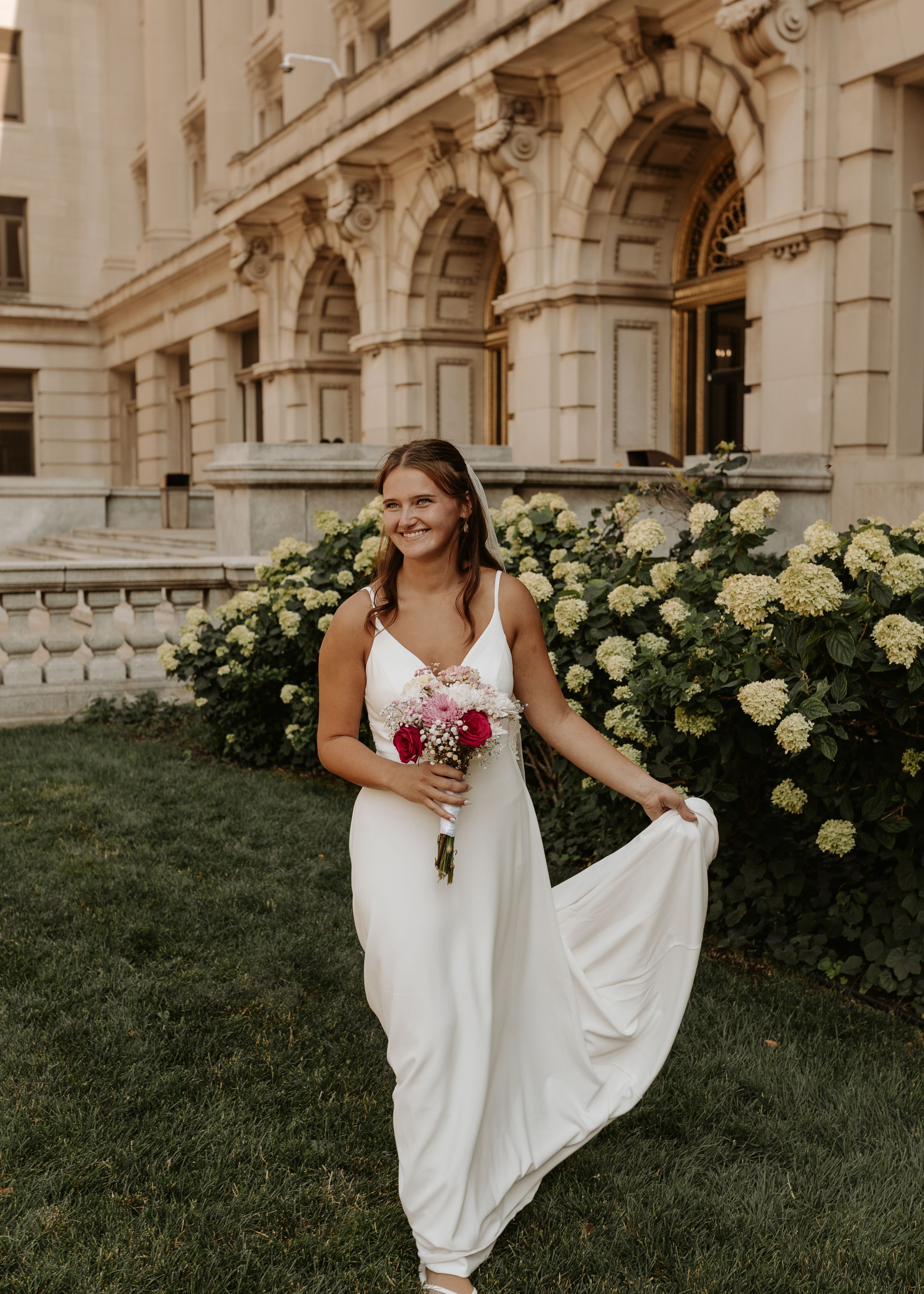 Bride in white gown holding pink bouquet smiles outside ornate stone building with hydrangea bushes.