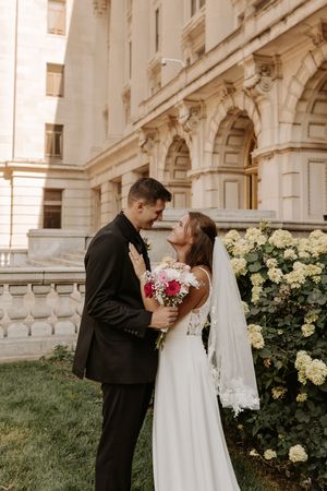 Bride and groom share a romantic moment outside a grand stone building, bride holding a pink and white bouquet.