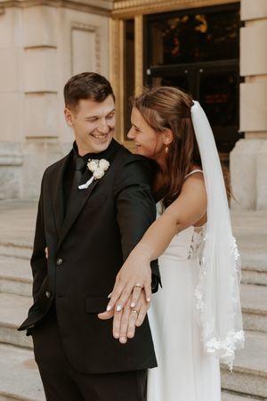 A smiling bride and groom in wedding attire display their rings outside a classic stone building.