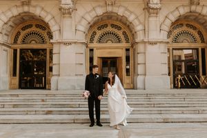 Bride and groom standing on steps of elegant stone building, bride in flowing white gown holding bouquet.