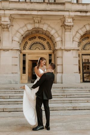 Groom lifting bride in white gown on steps of elegant stone building during outdoor wedding photo session.
