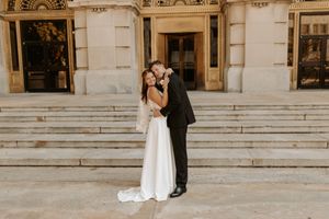 A bride and groom embrace and share a kiss on the steps of an elegant stone building on their wedding day.