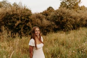 Young woman in white dress smiling in a sunlit meadow with lush green trees in the background.