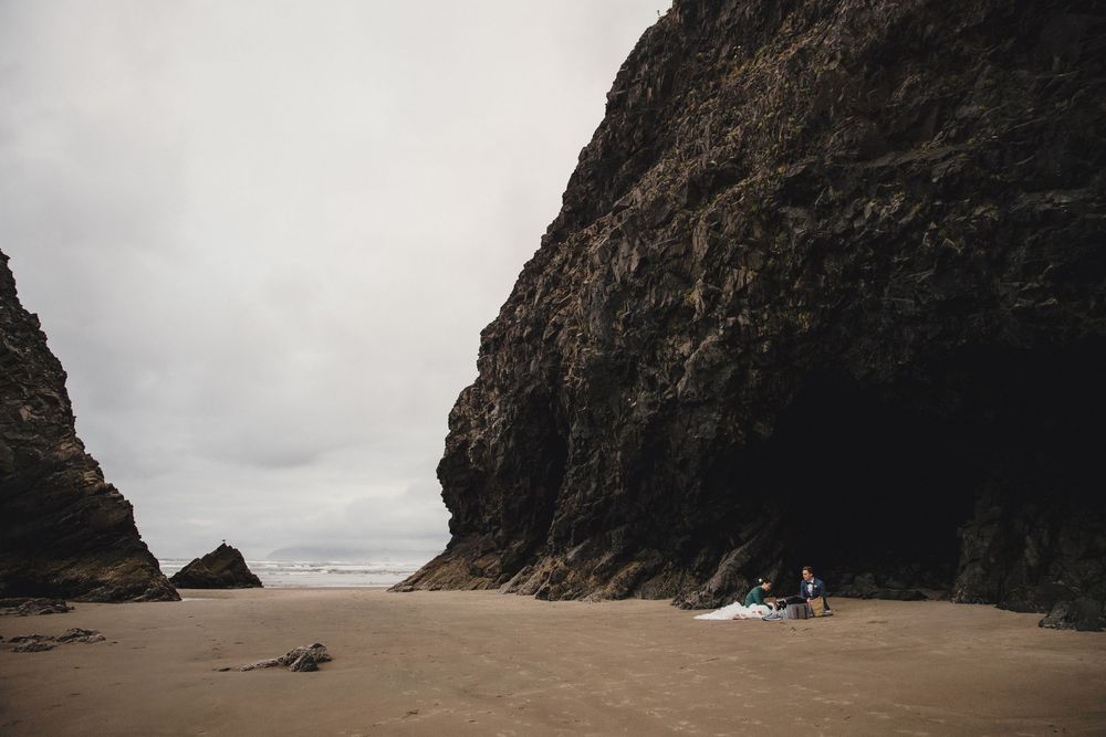 Couple sitting on a sandy beach beside towering dark rock cliffs under an overcast sky.