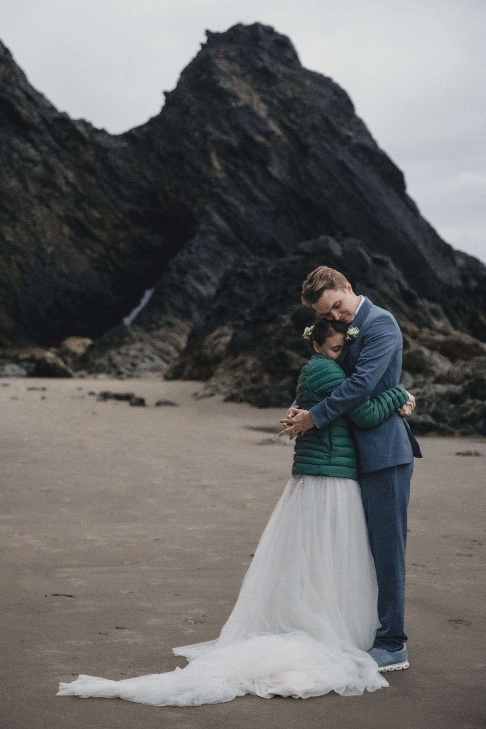 Bride and groom embrace on a sandy beach with dramatic rocky cliffs, bride wearing white skirt and green jacket.