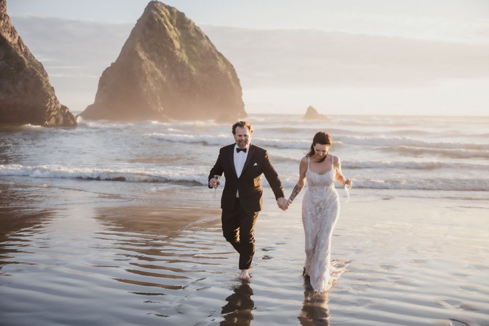 Bride and groom run hand-in-hand along a misty beach with dramatic sea stacks at golden hour.