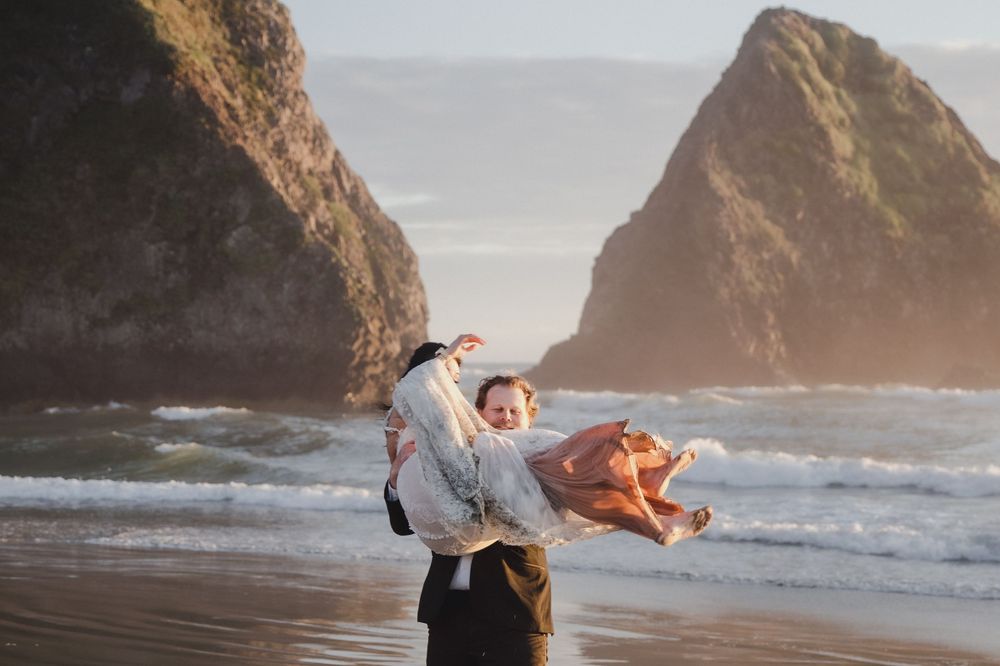 Person holding a baby on a scenic beach with dramatic rock formations and ocean waves in golden light.