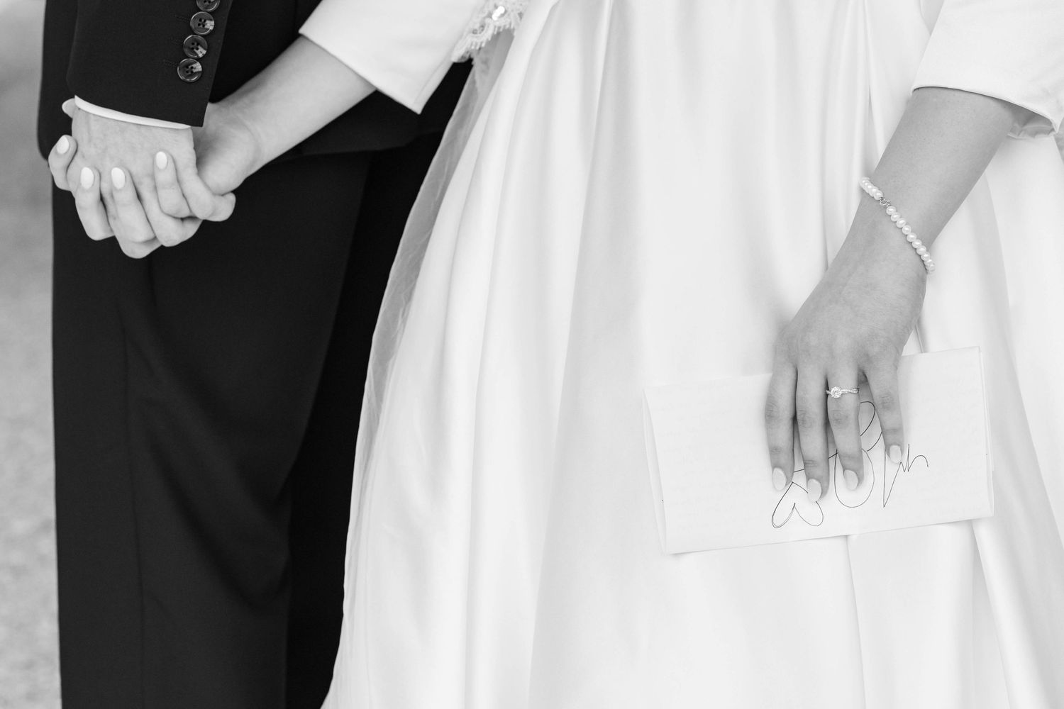 Black and white photo of bride and groom holding hands, showcasing wedding dress and suit at ceremony.