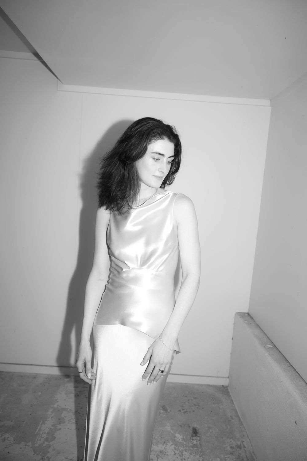 Woman in satin slip dress poses against white wall, black and white editorial fashion photography.