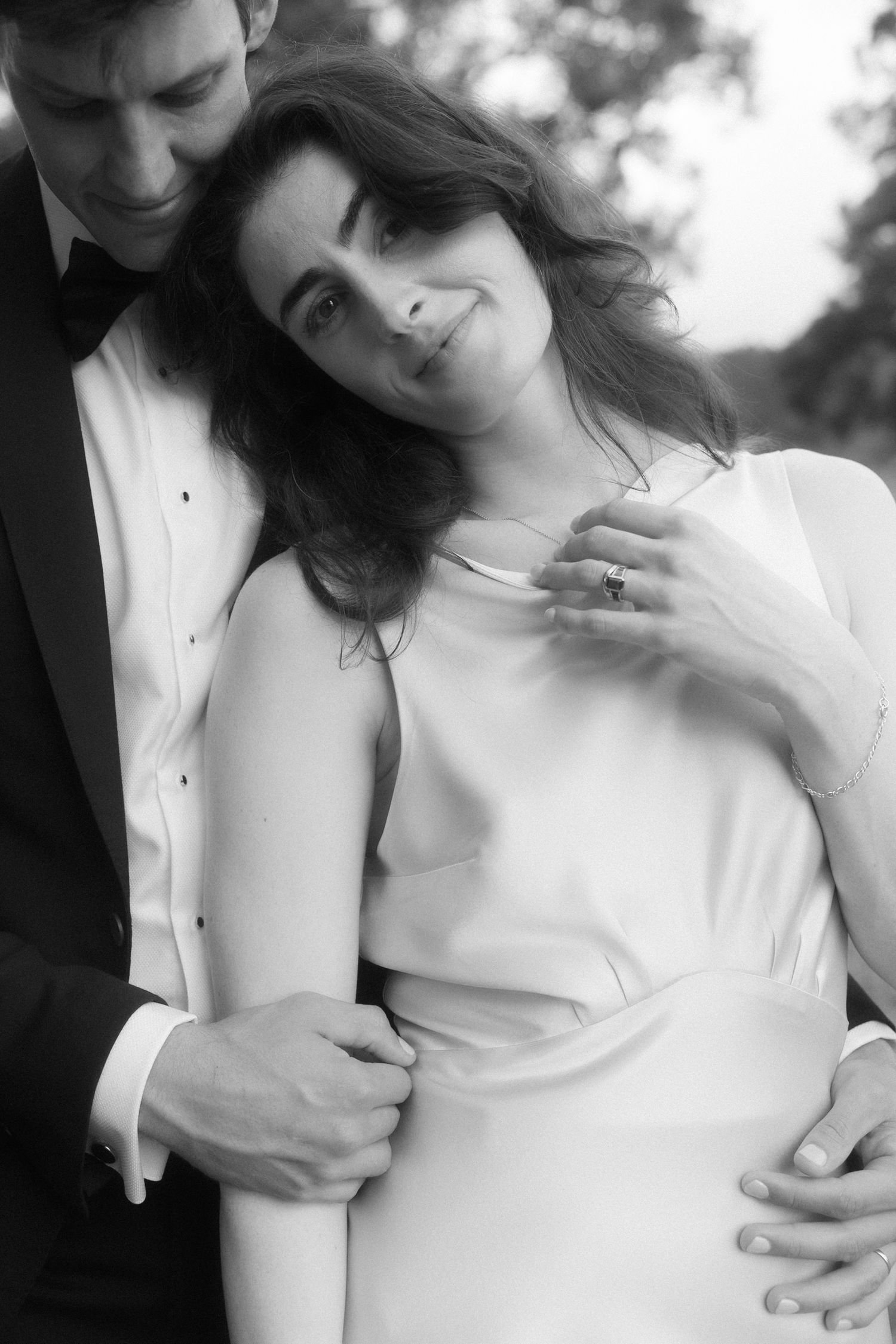 Black and white wedding photo of bride in white dress smiling as groom in tuxedo embraces her from behind outdoors.