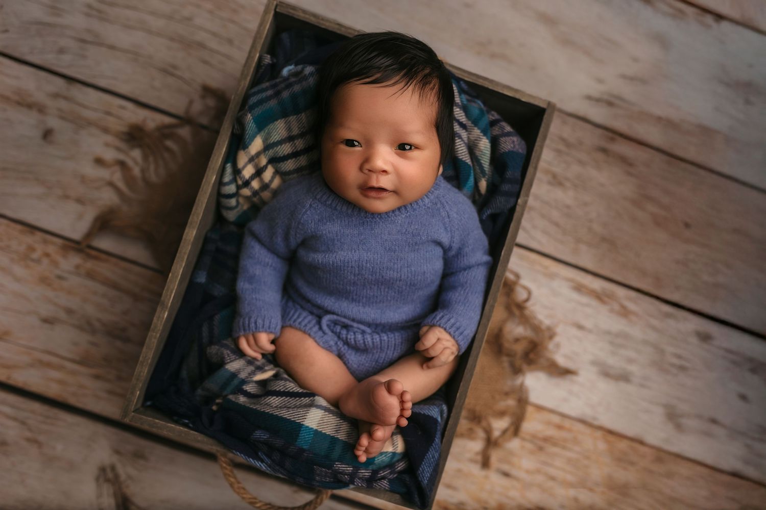 Newborn boy in a blue sweater smiles as he lays in a little crate against blue plaid, captured by Charlottesville baby photographer.