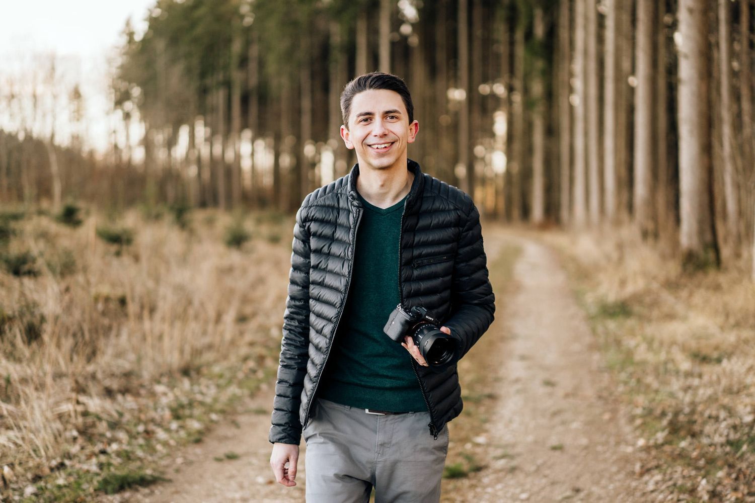 Young man in olive green t-shirt smiling outdoors against rustic brick wall background.