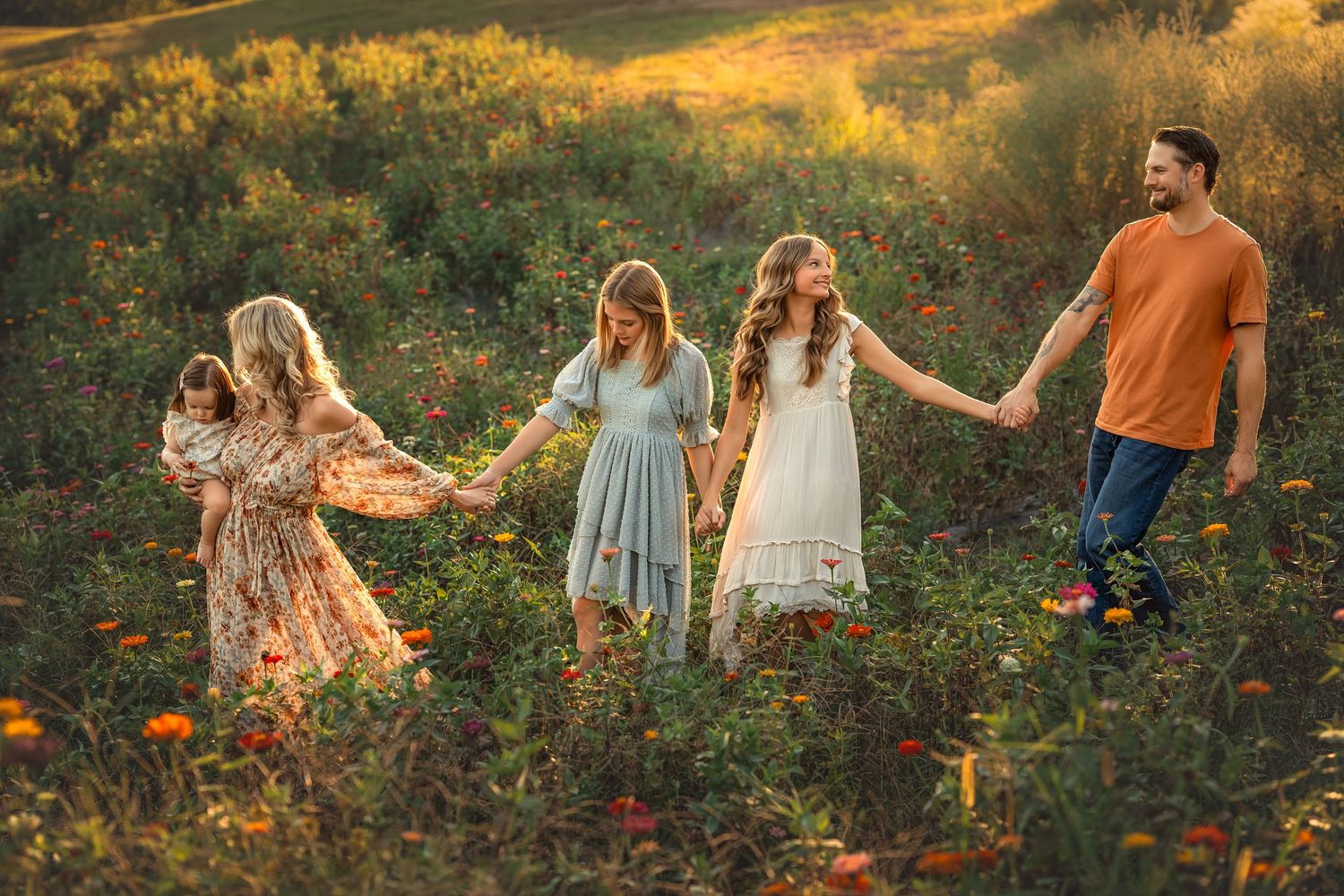 Family of five holding hands walking through a golden wildflower field at sunset in flowing dresses.