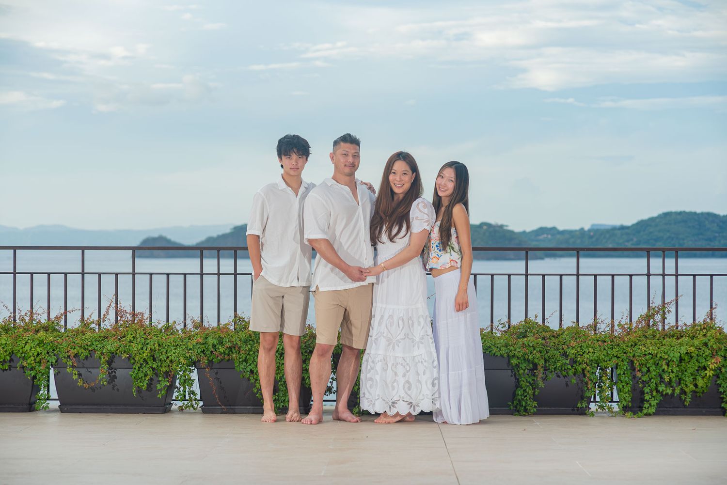 Family of four posing on a rooftop terrace overlooking a scenic ocean bay, dressed in white and neutral tones.