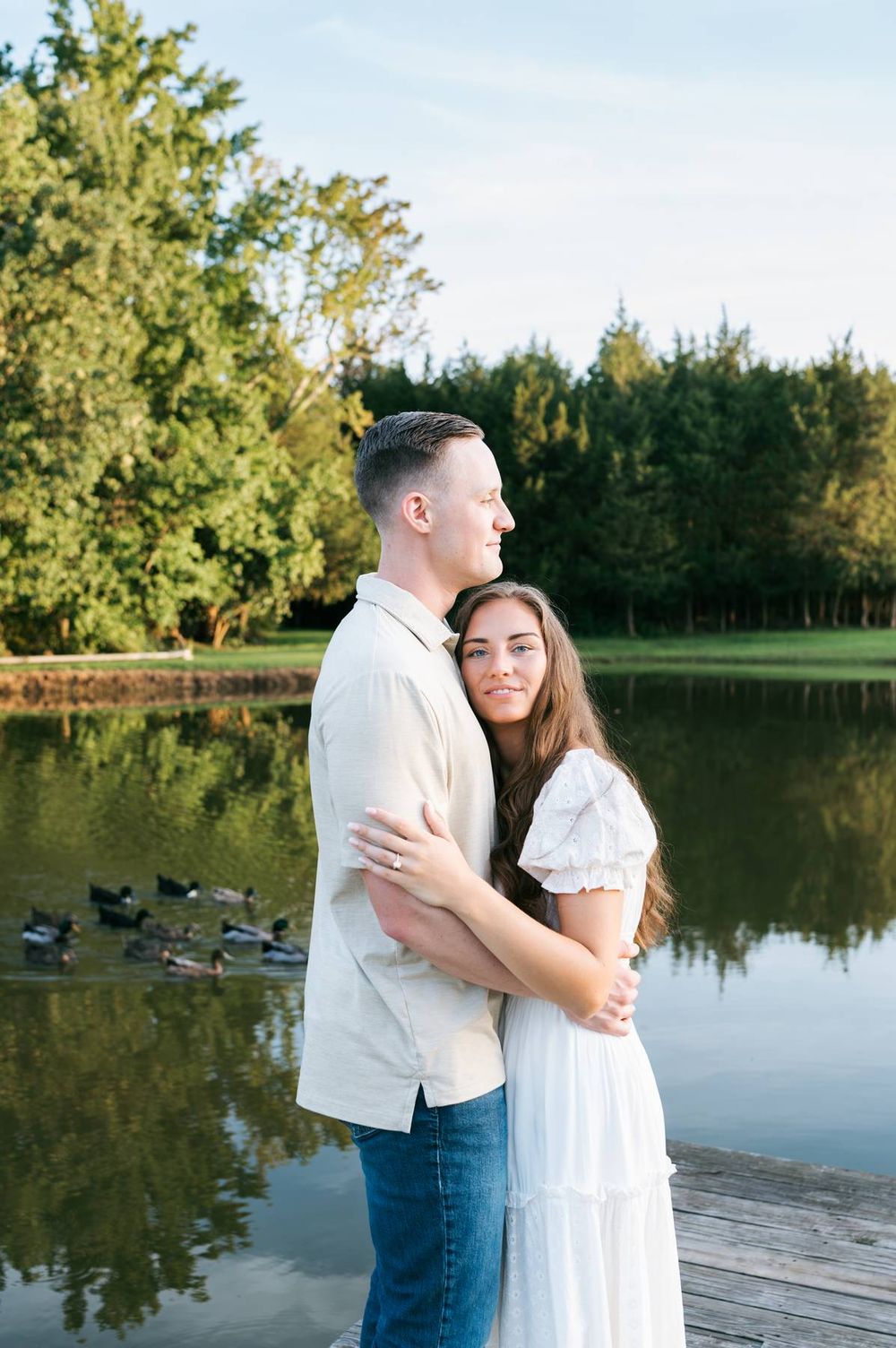 Engaged couple embracing on a wooden dock beside a serene pond surrounded by trees, engagement photographer Mooresville NC