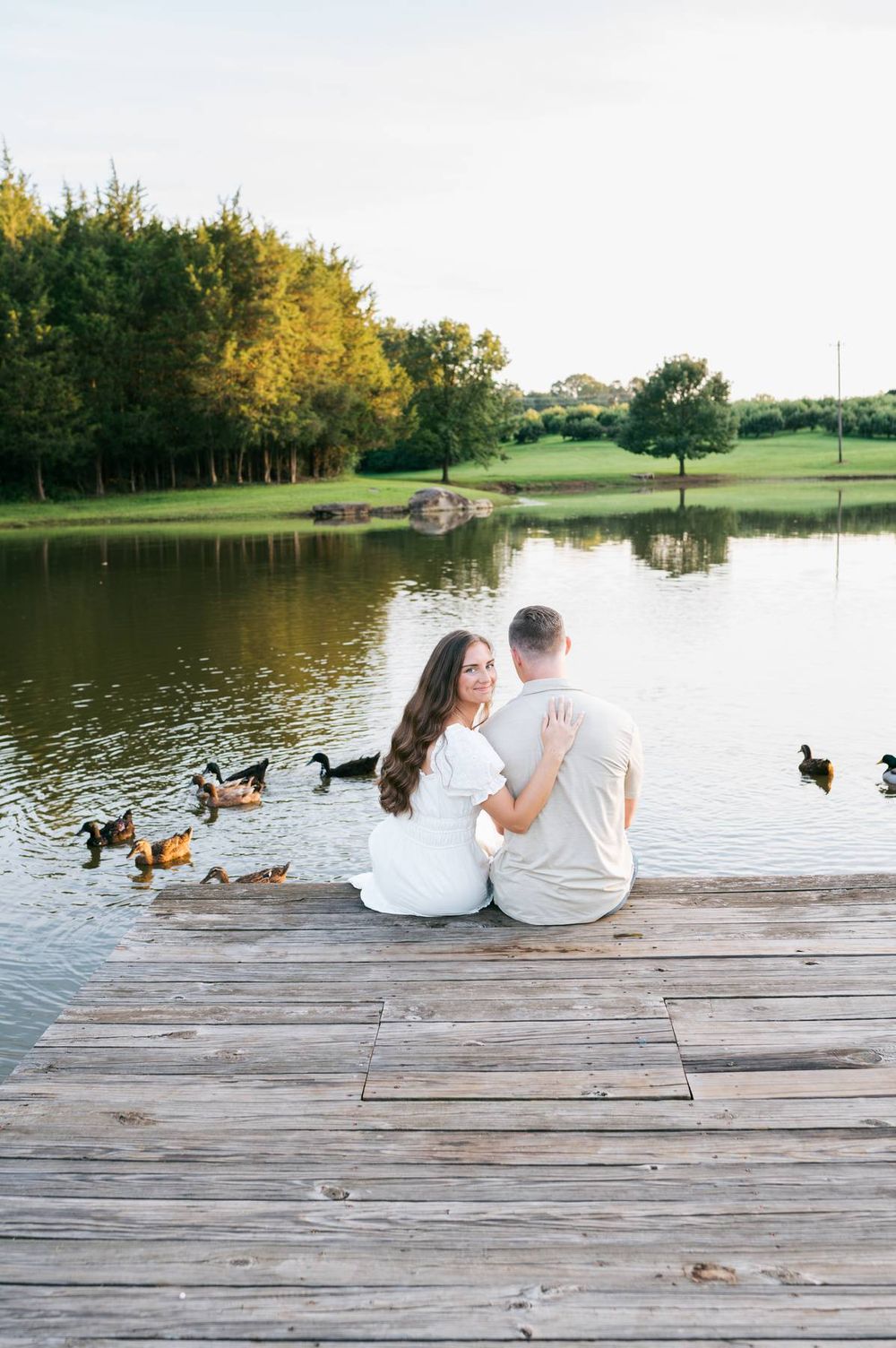 Couple sitting together on a wooden dock by a pond, surrounded by ducks at sunset, engagement photographer Mooresville NC